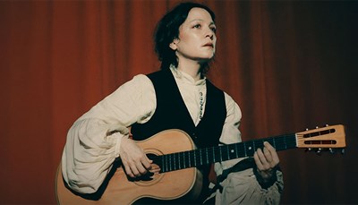 Natalia Lafourcade in a white blouse and black dress standing in front of a red curtain. Photo