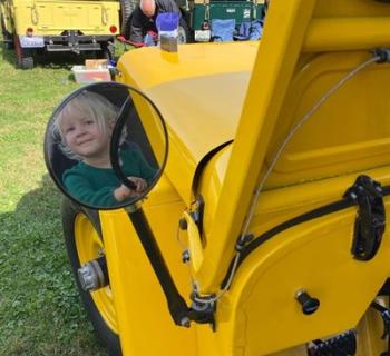 A toddler sits at the wheel of a historic jeep painted yellow. Photo