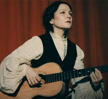 Natalia Lafourcade in a white blouse and black dress standing in front of a red curtain. Photo