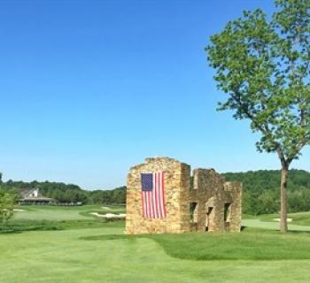 Picture of a fairway that has a stone wall with an American flag hanging along the side. Photo